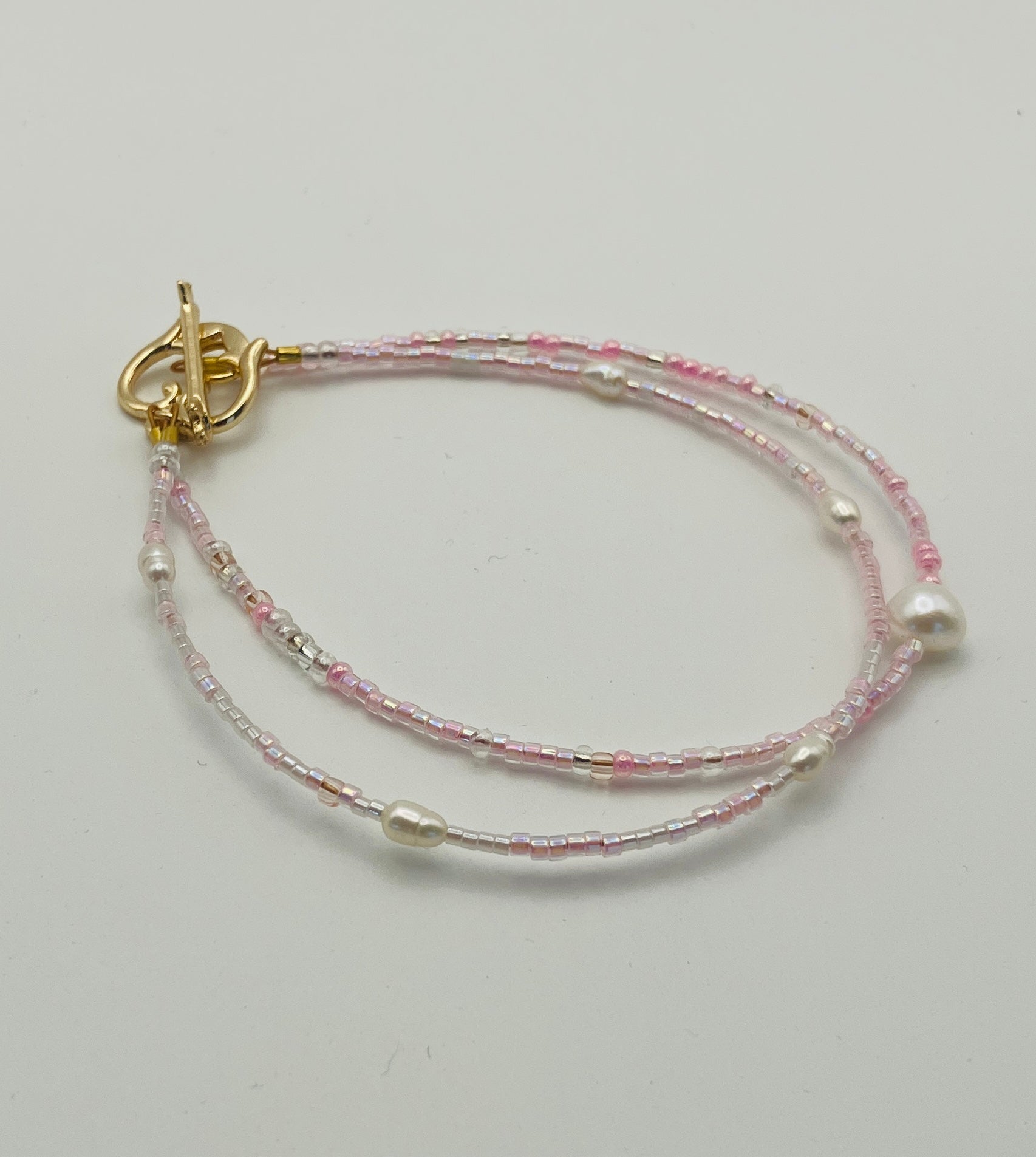 A delicate double-strand beaded bracelet with pink and silver beads, featuring irregular white pearls and a gold toggle clasp, shown on a plain light background.